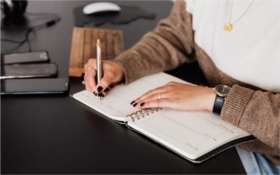 : Woman writing in a weekly planner at a dark wood desk, reviewing her schedule.
