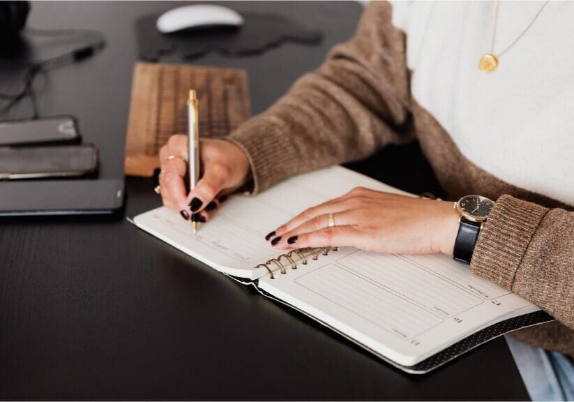 : Woman writing in a weekly planner at a dark wood desk, reviewing her schedule.